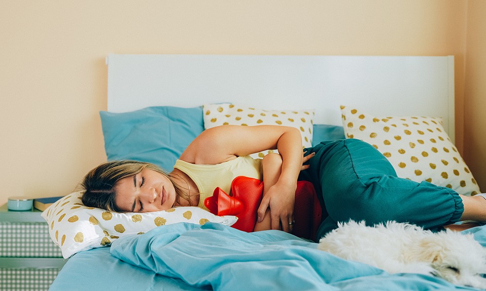 Frau liegt schmerzerfüllt mit Wärmflasche in ihrem Bett 