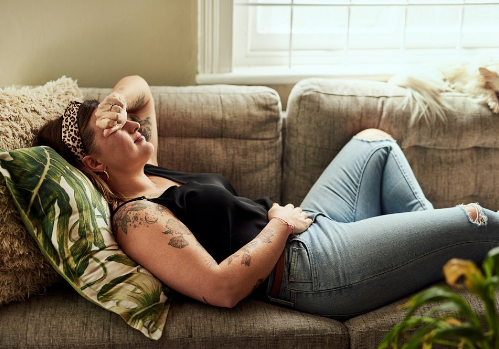Frau liegt mit Unterleibsschmerzen auf der Couch - Eine Hand lehnt auf dem Kopf, die zweite Hand liegt auf ihrem Unterleib
