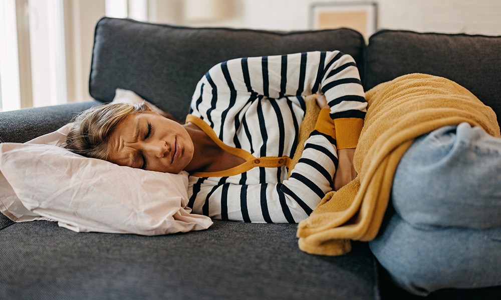 Frau liegt schmerzerfüllt mit einer Decke in Embryostellung auf der Couch 